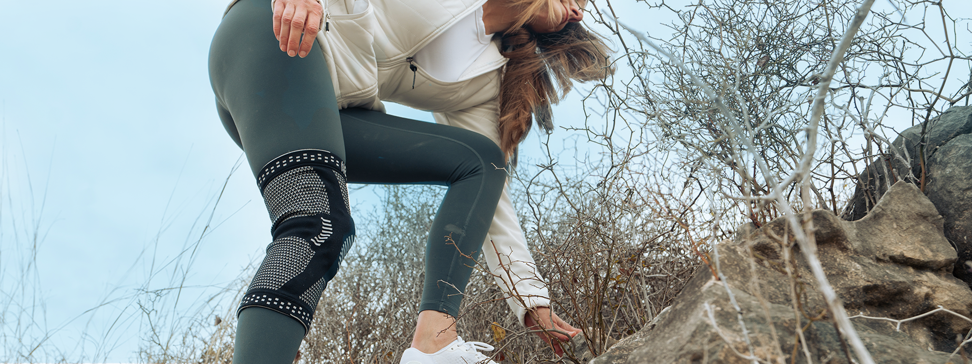 A woman in a white long-sleeve top and dark gray leggings with black knit knee panels is crouching outdoors among dry branches and rocks under a pale blue sky.