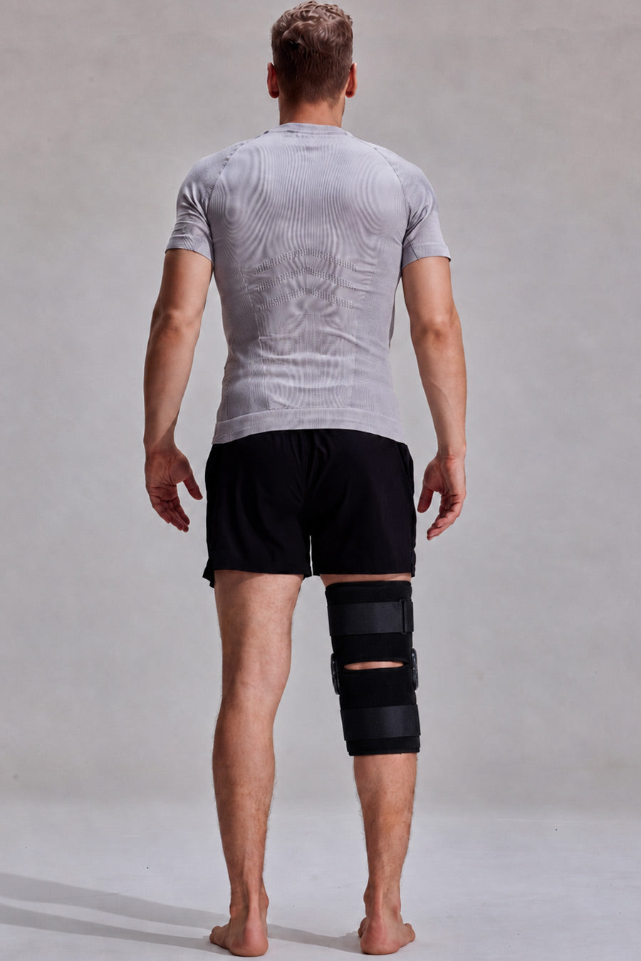 A man viewed from behind wearing a light gray textured t-shirt, black athletic shorts, and a black knee immobilizer brace on his right leg, standing against a neutral light gray studio background.