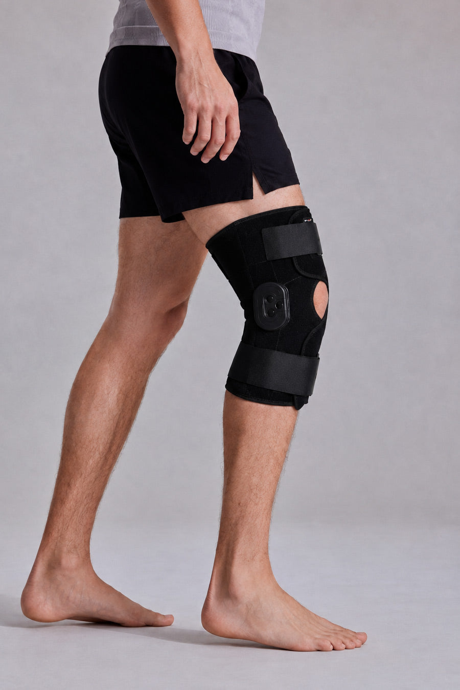 A man wearing ace-49 knee immobilizer brace on his right leg, paired with black athletic shorts, shown from the waist down against a neutral gray studio background.
