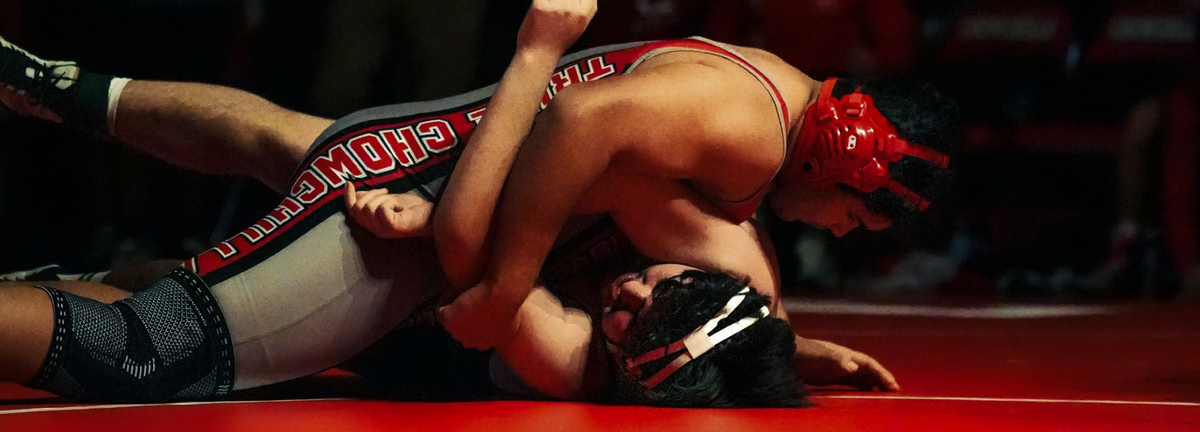 Two wrestlers in action on a red mat with one wrestler in a red and white uniform being held by another.