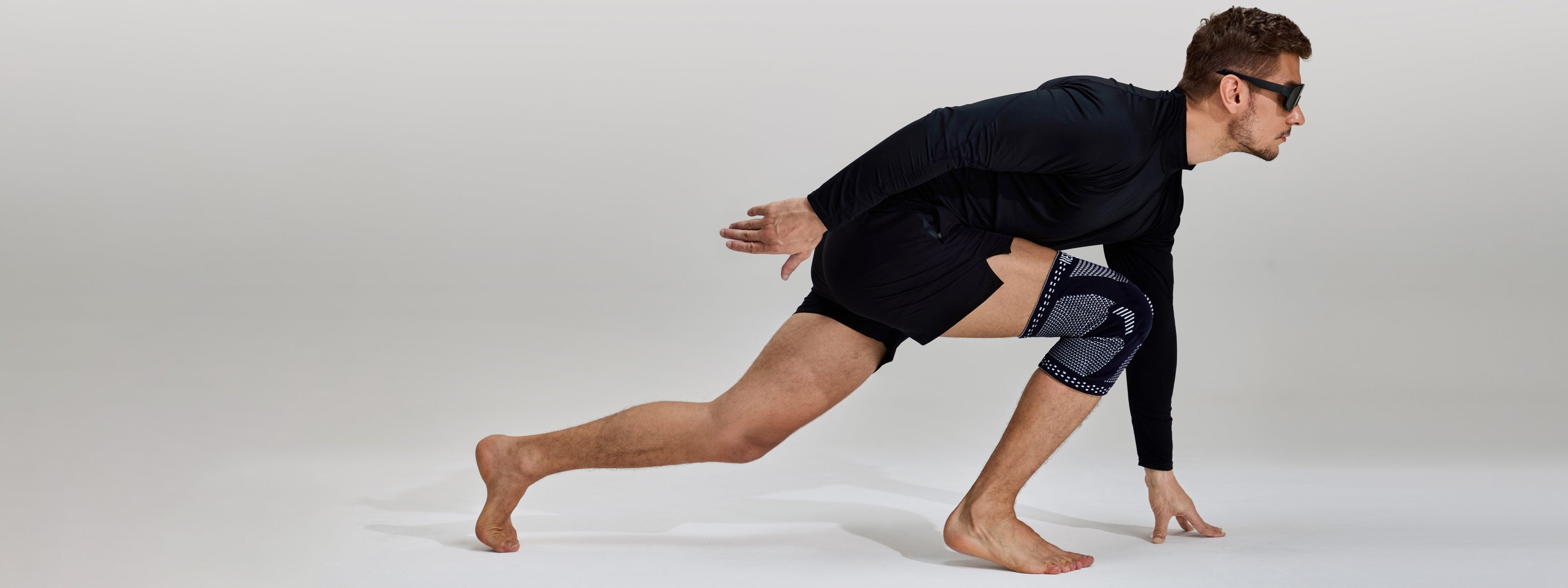 A man in a black long-sleeve top and black athletic shorts is crouching in a low athletic pose on a light gray studio background. He is wearing a dark knee support brace on his right leg and sunglasses.