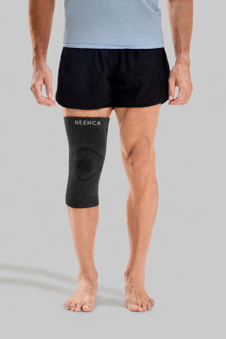 A man wearing a black Neenca knee support sleeve on his right leg, paired with black athletic shorts and a light blue t-shirt, standing against a solid gray studio background.