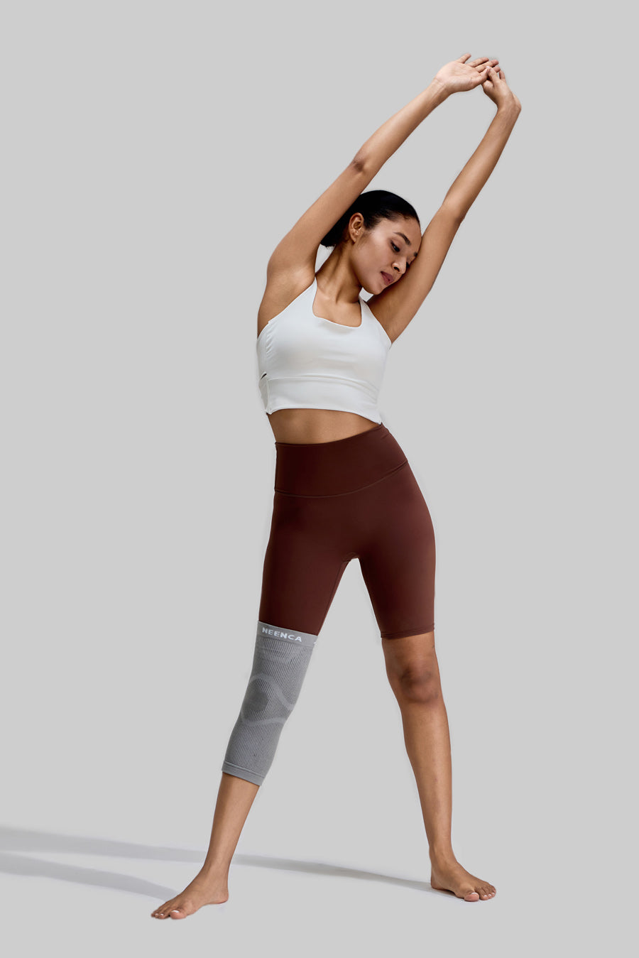 Female model performing an overhead side stretch while wearing the grey NEENCA HX038 knee brace on her right leg, showcasing the product's flexibility during exercise