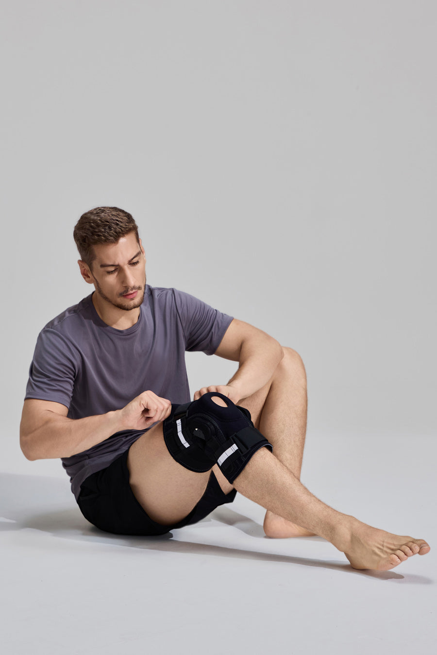 Male model sitting down while adjusting the compression straps of a NEENCA professional hinged knee brace HX043 for a personalized fit