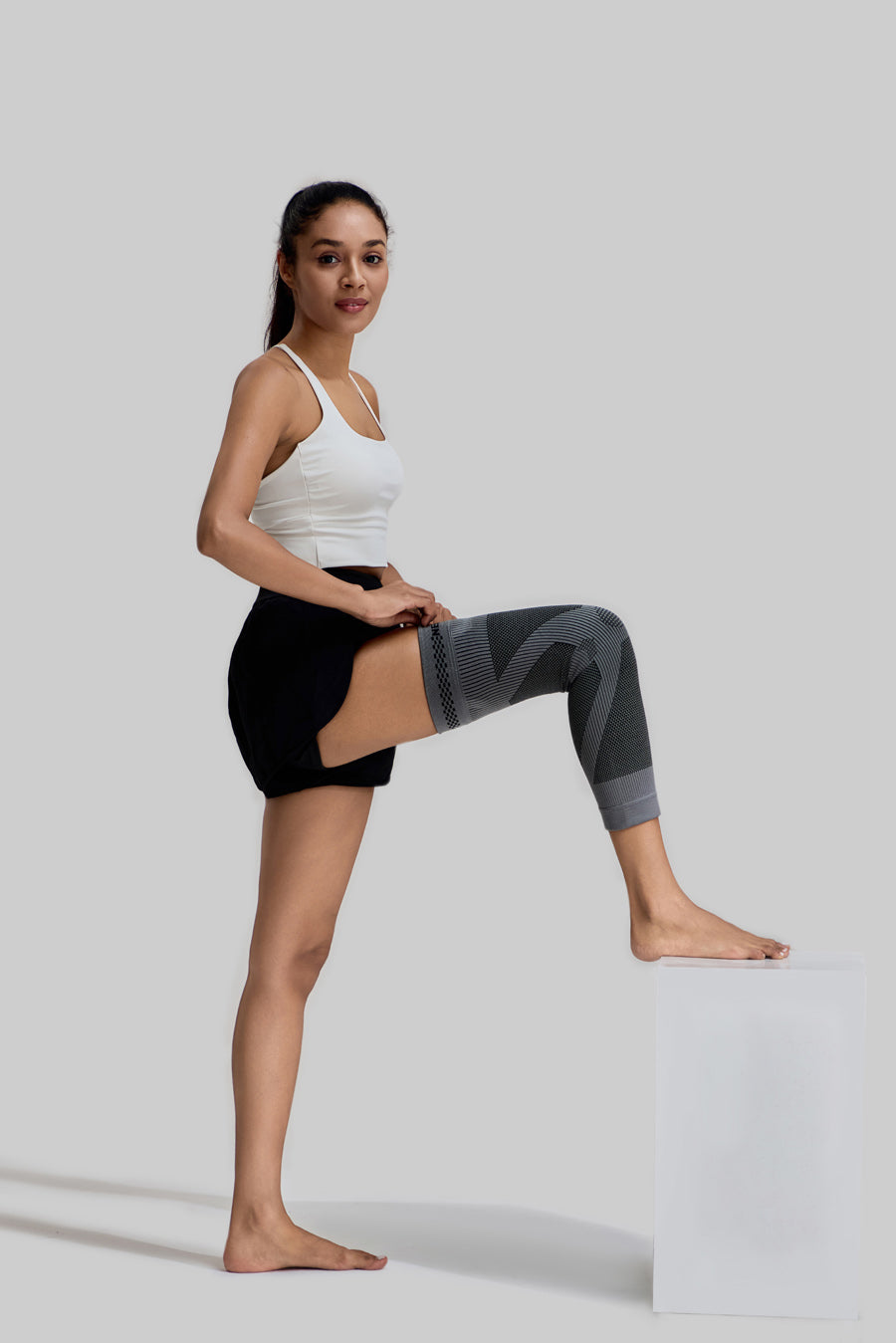 Side view of a female model adjusting the grey NEENCA HX036 leg sleeve with her foot resting on a white stool