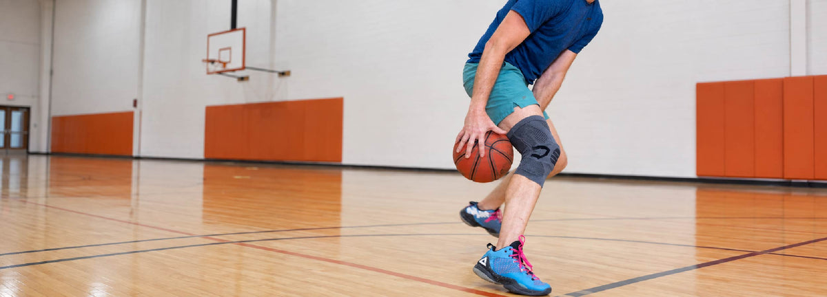 A basketball player dribbling on an indoor court while wearing a gray NEENCA knee brace, highlighting knee support during quick movement and direction changes.