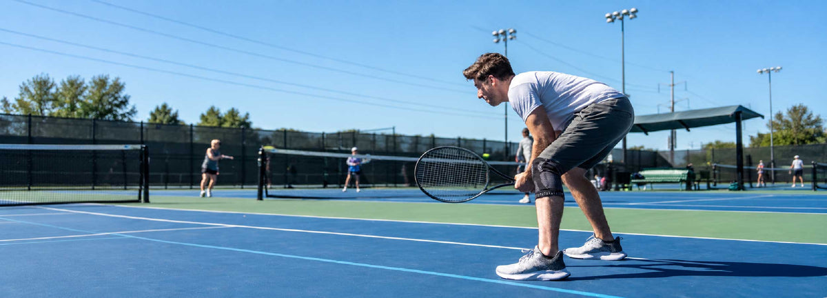 A tennis player wearing a NEENCA knee brace on an outdoor court, preparing for lateral movement and quick directional changes during play.