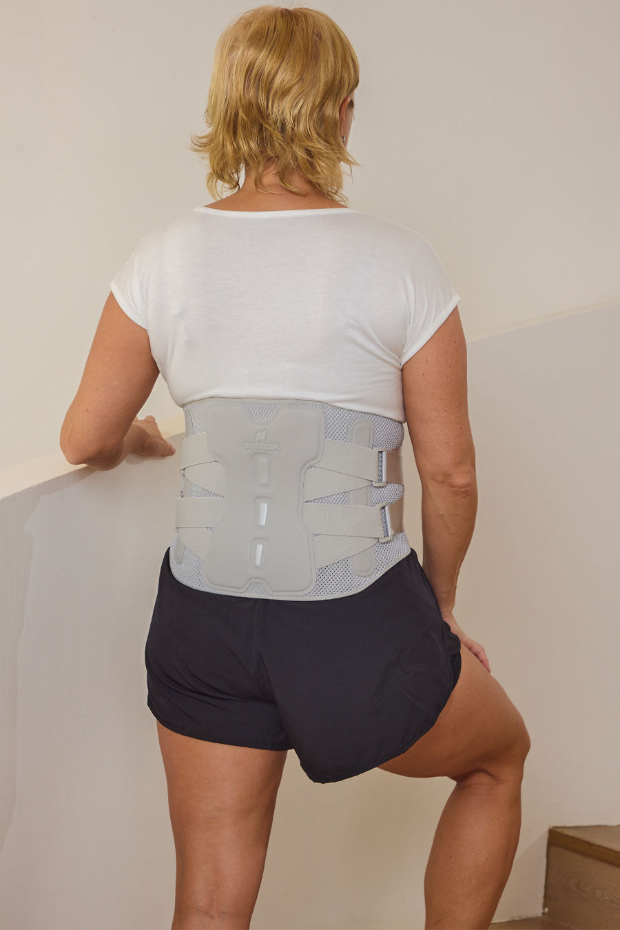 A woman viewed from behind wearing a light gray back support brace over a white t-shirt and dark shorts, standing indoors against a plain wall.