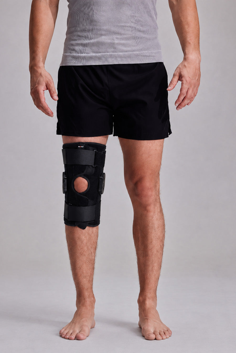 A man wearing ace-49 knee immobilizer brace on his right leg, paired with black athletic shorts and a gray t-shirt, standing against a light gray studio background.