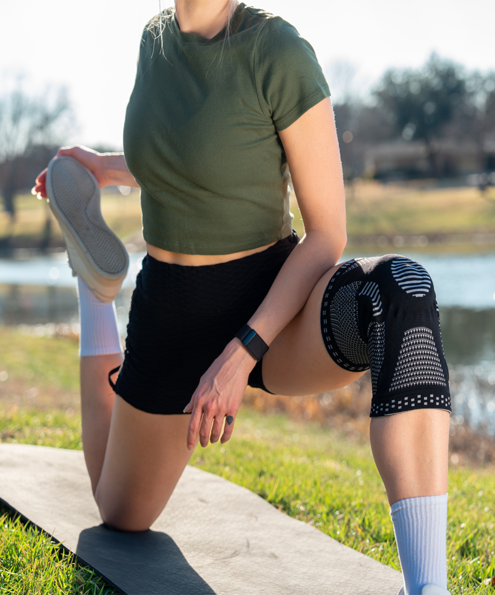 Person stretching outdoors while wearing Neenca knee braces on a grassy area with a lake in the background