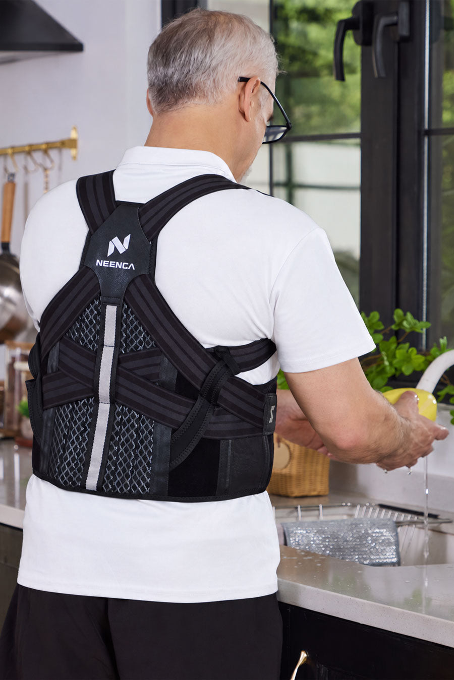 A man viewed from behind in a kitchen, wearing a black posture corrector brace with a white NEENCA logo on the upper back panel.