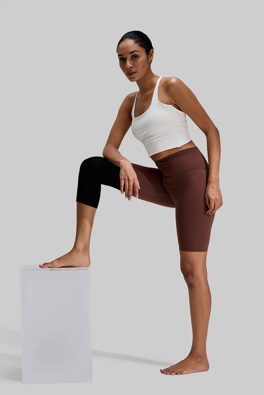 Full-body view of a female model wearing the black NEENCA HX038 knee brace on her right leg, with her foot resting on a white stool to demonstrate the brace's flexible and supportive fit during exercise