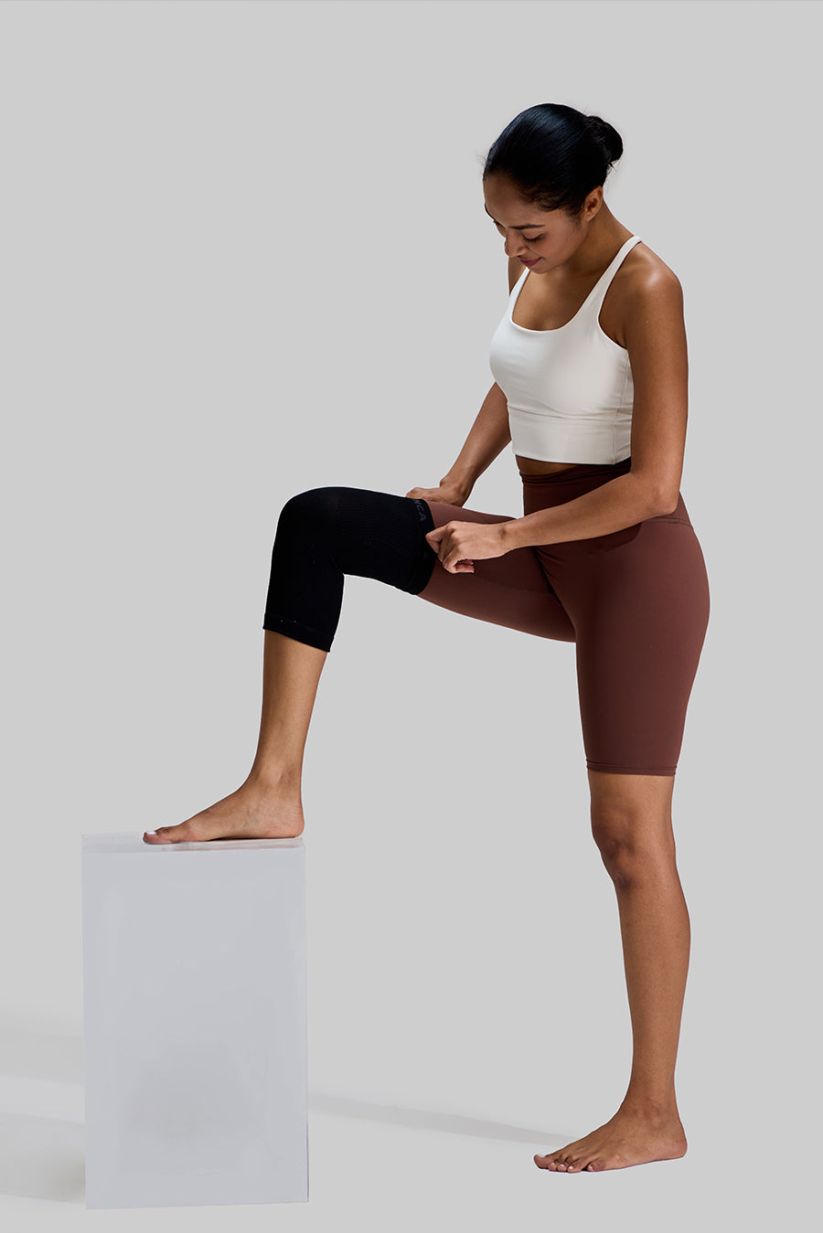 Female model adjusting the black NEENCA HX038 knee brace on her right leg, with her foot resting on a white stool to demonstrate the ease of application and secure fit