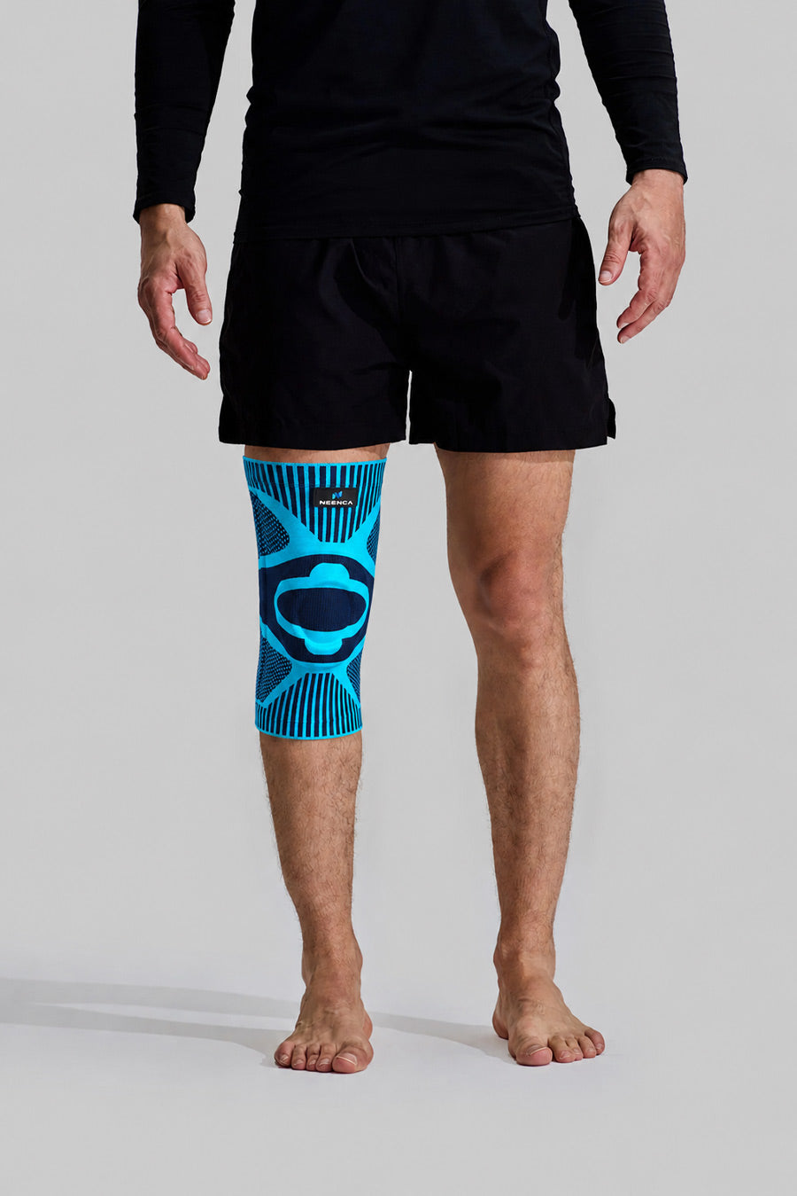 A man wearing black athletic shorts and a black long-sleeve top, standing barefoot against a light gray studio background. His right leg is fitted with a neenca ace-53 blue knee compression sleeve featuring a dark abstract graphic.